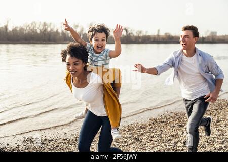 Padre che corre dietro la madre piggybacking figlio mentre corre da Lakeshore Foto Stock