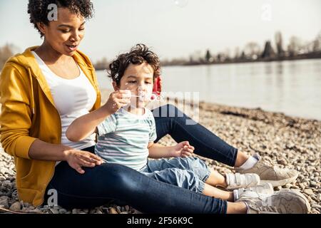 Ragazzo che soffia nella bacchetta della bolla mentre si siede con la donna ciottoli Foto Stock
