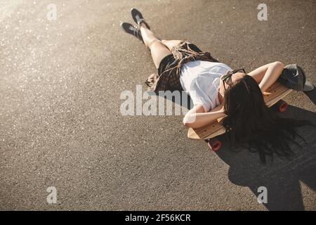 Stile di vita dello skater, concetto di svago urbano e di gioventù. Foto posteriore superiore hipster ragazza adolescente godere di vacanze estive, sdraiato su strada in cemento di skatepark on Foto Stock