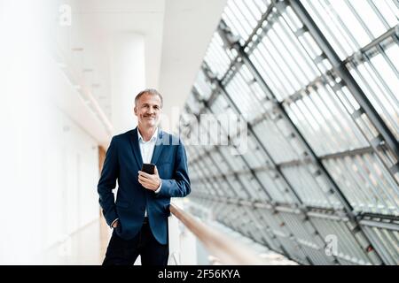 Sorridente bell'uomo d'affari che tiene il telefono cellulare mentre si sta in piedi alla ringhiera nel corridoio degli uffici Foto Stock