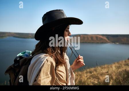 Vista laterale della bella donna nel cappello da cowboy con zaino che passa il tempo libero in aria fresca. Attraente viaggiatore escursioni da solo al parco nazionale Podillya Tovtry. Foto Stock