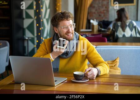 Imprenditore maschile con portatile che invia messaggi vocali tramite telefono cellulare mentre si ha un caffè al caffè Foto Stock