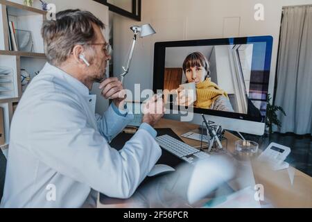 Medico maturo che parla con il paziente in videochiamata su computer mentre sedendo all'ufficio del medico Foto Stock