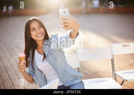 Giovane donna che fa selfie con gelato su un palcoscenico in un parco cittadino in una giornata luminosa e soleggiata. Delizioso Foto Stock