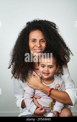 Donna sorridente con capelli ricci che porta figlia mentre si siede contro il grigio sfondo Foto Stock