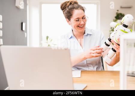 Felice ingegnere donna che tiene il robot mentre si siede alla scrivania Foto Stock