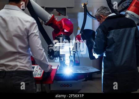 Saldatore maschio che esamina la robotica mentre si trova in piedi da un giovane ingegnere che lavora in fabbrica Foto Stock