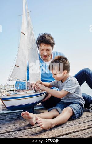 Ragazzo legando corda di barca giocattolo mentre si siede con l'uomo sul molo Foto Stock