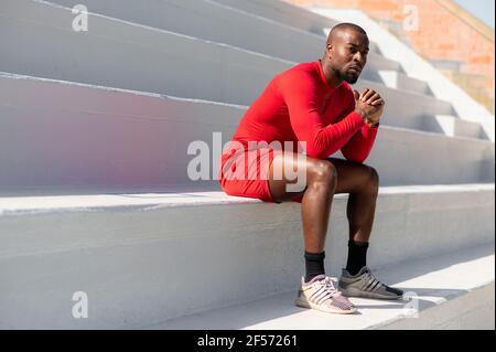 Vista laterale sportivo nero afroamericano seduto su scale bianche in uno stadio con mani unite che guardano lontano con un volto serio e mirato. Foto Stock