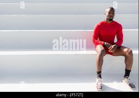 Uomo muscoloso africano nero con abbigliamento sportivo seduto su scalini bianchi sulla scala e guardando lontano. Minimalismo con spazio di copia Foto Stock