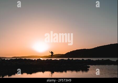 Profilo grandangolare del viaggiatore in riva al mare che salta in spiaggia durante il tramonto. Foto Stock