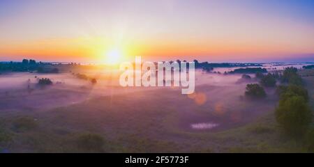 Splendido paesaggio rurale con alberi colorati e foggosi, cielo arancione con sole. Vista aerea del villaggio in nebbia con raggi solari dorati all'alba Foto Stock