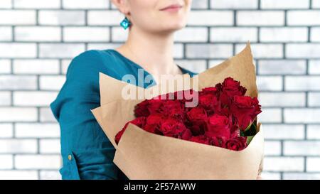 Una bella ragazza in una camicia blu contiene un bouquet di rose rosse di lusso avvolto in carta artigianale e sorrisi. Combinazione di colori rosso e blu. Fiori come Foto Stock
