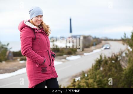 Donna vestendo caldo cappotto rosso in piedi sulla collina contro il faro sul capo mare, viaggio nella stagione invernale Foto Stock