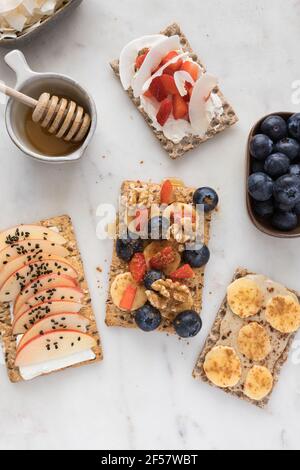 Vista dall'alto di quattro deliziosi cracker con frutta, noci e spezie sopra di loro accanto a una ciotola piena di miele con un cucchiaino di legno all'interno su un bo di marmo Foto Stock