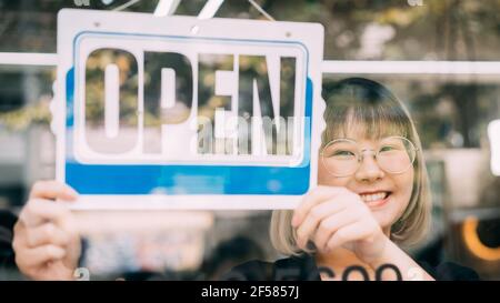 Giovane donna asiatica appesa al segno aperto dalla finestra di vetro. Proprietario del negozio femminile con avviso aperto davanti al negozio Foto Stock