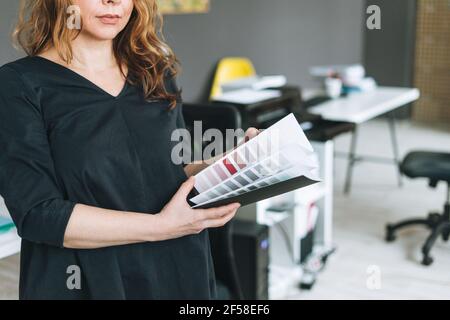 Affascinante donna adulta di mezza età con capelli ricci architetto designer con palette di colori in mano in un moderno momento di lavoro in ufficio Foto Stock