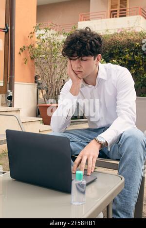 Un giovane di 18 anni, con la mano a faccia, con espressione di frustrazione, sta guardando il suo laptop, seduto fuori. Gel disinfettante sul tavolo. Foto Stock