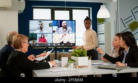 Black manager donna che parla con colleghi in remoto in videochiamata sullo schermo tv, presentando nuovi partner commerciali. Donna d'affari sicura che parla con la webcam che trasmette formazione del webcast, che fa la chiamata in conferenza in linea Foto Stock