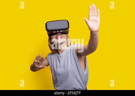 La donna bionda caucasica sta toccando qualcosa mentre sperimenta un nuovo aumento reality headset su una parete gialla dello studio Foto Stock