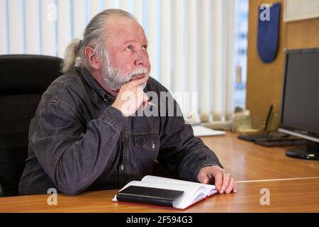 Pensare caucasico senior business persona seduta alla scrivania in sala ufficio con penna e organizer notebook Foto Stock