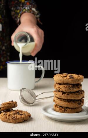 Primo piano di una pila di biscotti al cioccolato su piatto bianco e la mano della donna che serve il latte in tazza bianca, fuoco selettivo, su tavolo di legno bianco, sfondo nero Foto Stock