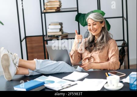 Bella fiduciosa influente donna asiatica con capelli grigi maturi, manager, leader aziendale, freelancer che chiacchiera con i dipendenti tramite collegamento video, gettando i piedi sul tavolo, approvazione gesturing, sorridendo Foto Stock