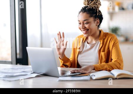 Comunicazione video online. Gioiosa attraente donna afro-americana, freelancer o studente, in abiti casual, usa il laptop per videoconferenza online con colleghi o amici, saluta, sorrisi Foto Stock