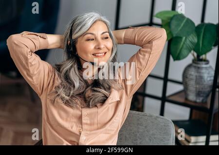 Gioiosa donna d'affari asiatica con capelli grigi, top manager, freelance, seduta in ufficio, facendo una pausa durante il lavoro, incrociando le braccia dietro la testa, sorridendo Foto Stock