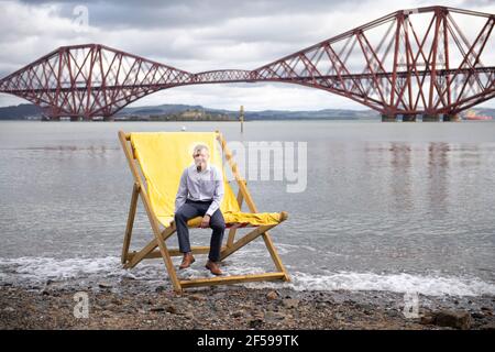 South Queensferry, Scozia, Regno Unito. 25 Marzo 2021. NELLA FOTO: Willie Rennie MSP. Willie Rennie MSP - leader del Partito Liberale democratico Scozzese (Scottish Lib Dems) si è Unito a Rebecca Bell, candidato di Edimburgo Northern e Leith, e a sua figlia Daphne. Legge la figlia di Rebecca un libro su una sedia gigante da spiaggia con la vista del Forth Bridge dietro come parte del loro percorso di campagna elettorale Holyrood per il 6 maggio. Credit: Colin Fisher/Alamy Live News Foto Stock