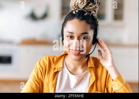 Primo piano di afroamericano una giovane donna attraente e fiduciosa in cuffia, seduta in ufficio, che lavora come operatore di call center o servizio di assistenza, guarda direttamente la fotocamera e sorridente amichevole Foto Stock