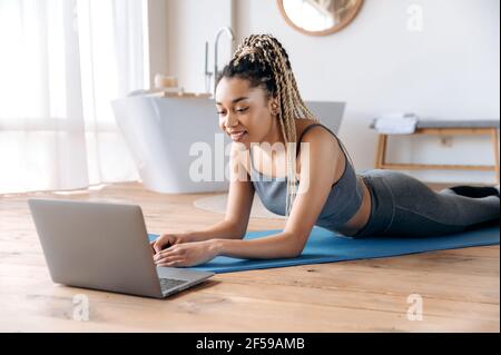 Felice atletica giovane afroamericana donna con una buona figura, in un vestito sportivo, si trova sul pavimento a casa, guarda online lezioni di fitness da portatile, entra per lo sport a casa, conduce uno stile di vita sano Foto Stock