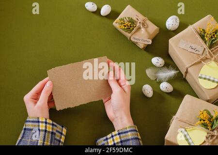 Mani femminili che tengono una carta bianca fatta a mano, scatole di regalo di pasqua decorate con fiori, un'etichetta a forma di uovo. Nessun regalo di spreco. Foto Stock