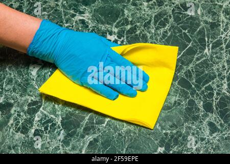 La mano della casalinga in un guanto di lattice blu pulisce una superficie di marmo verde con un tovagliolo giallo in cucina. Foto Stock