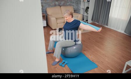 Donna anziana che siede sull'addestramento della sfera di equilibrio con la banda di resistenza. Allenamento di anziani a casa sport stile di vita sano, allenamento di esercizi di fitness anziani in appartamento, attività e assistenza sanitaria Foto Stock