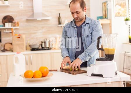 Guy prepara frullato nutriente in cucina con frutta fresca. Stile di vita sano e spensierato, dieta e preparazione della colazione in un'accogliente mattina di sole Foto Stock