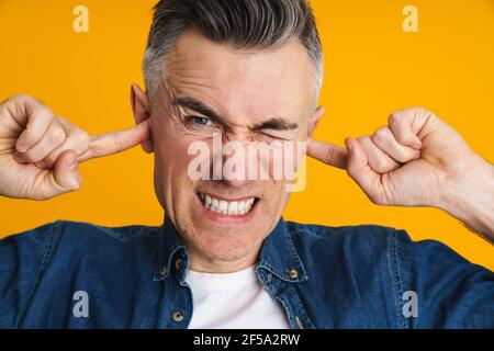 Dispiaciuto uomo bel grimacing mentre tappava le orecchie isolato sopra sfondo giallo Foto Stock