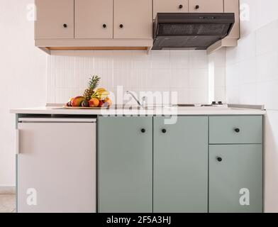 Closeup di piastrelle bianche splashback su parete vuota in semplice armadio modulo di moderna cucina interna con piccolo frigorifero, sfiato, lavello, piano cottura, piatto di frutta Foto Stock