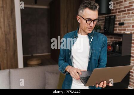 Uomo bello messo a fuoco negli occhiali usando gli auricolari ed il laptop a. casa Foto Stock