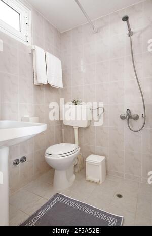 Interni in stile semplice di piccoli servizi igienici con pareti in piastrelle di ceramica beige, lavabo bianco, wc classico, tappeto ornamentale di meandro grigio, doccia aperta senza doccia Foto Stock