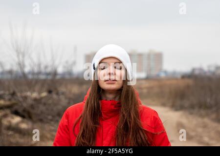 Bella giovane donna con capelli lunghi inala aria fresca con i suoi occhi chiusi in un cappello bianco lavorato a maglia e un giacca rossa con cappuccio sulla strada Foto Stock