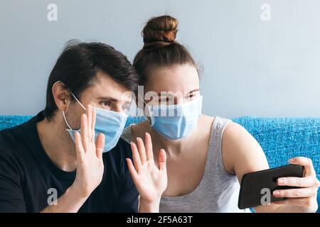 Una coppia sorridente in maschere mediche blu accoglie i genitori o gli amici in una chiamata online tramite smartphone Foto Stock