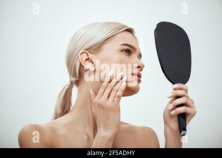 Perfezione. Profilo ritratto di giovane donna attraente che tocca il viso e guarda in piccolo specchio mentre si trova in piedi su sfondo grigio Foto Stock