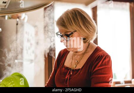 Donna anziana che cucina pranzo in cucina - donna matura in cucina drenando la pasta dall'acqua bollente - Boomer donna occupato cucina pranzo f Foto Stock