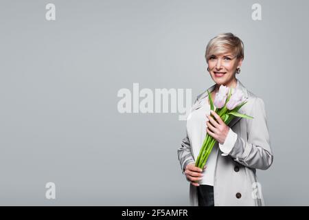 donna trendy di mezza età che sorride alla macchina fotografica mentre tiene i tulipani freschi isolati sul grigio Foto Stock