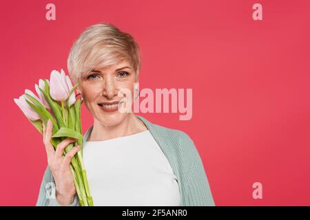 donna elegante che sorride alla macchina fotografica mentre tiene i tulipani freschi isolati su rosa Foto Stock