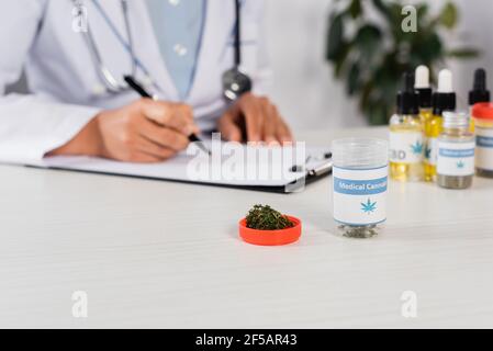 bottiglia con scritta sulla cannabis medica e erbacce verdi essiccate vicino medico che scrive prescrizione su sfondo offuscato Foto Stock