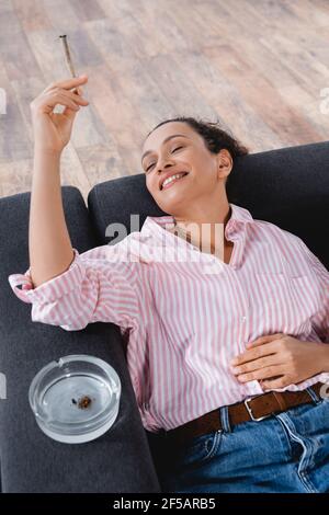 vista ad alto angolo di una donna afro-americana che si sdraiava divano e fumo unito con cannabis medica vicino posacenere Foto Stock