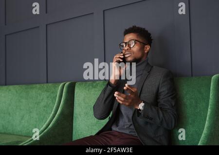 Giovane africano che parla con il suo collega sul telefono cellulare seduta su un bellissimo divano verde Foto Stock