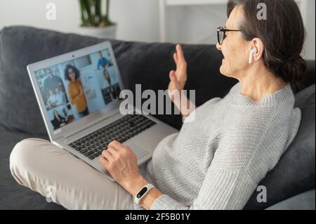 Donna moderna di pensione salutare i partecipanti alla riunione on-line, ha videoconferenza con i colleghi a distanza, donna anziana sdraiata sul divano, utilizzando il laptop per videochiamare team diversi Foto Stock